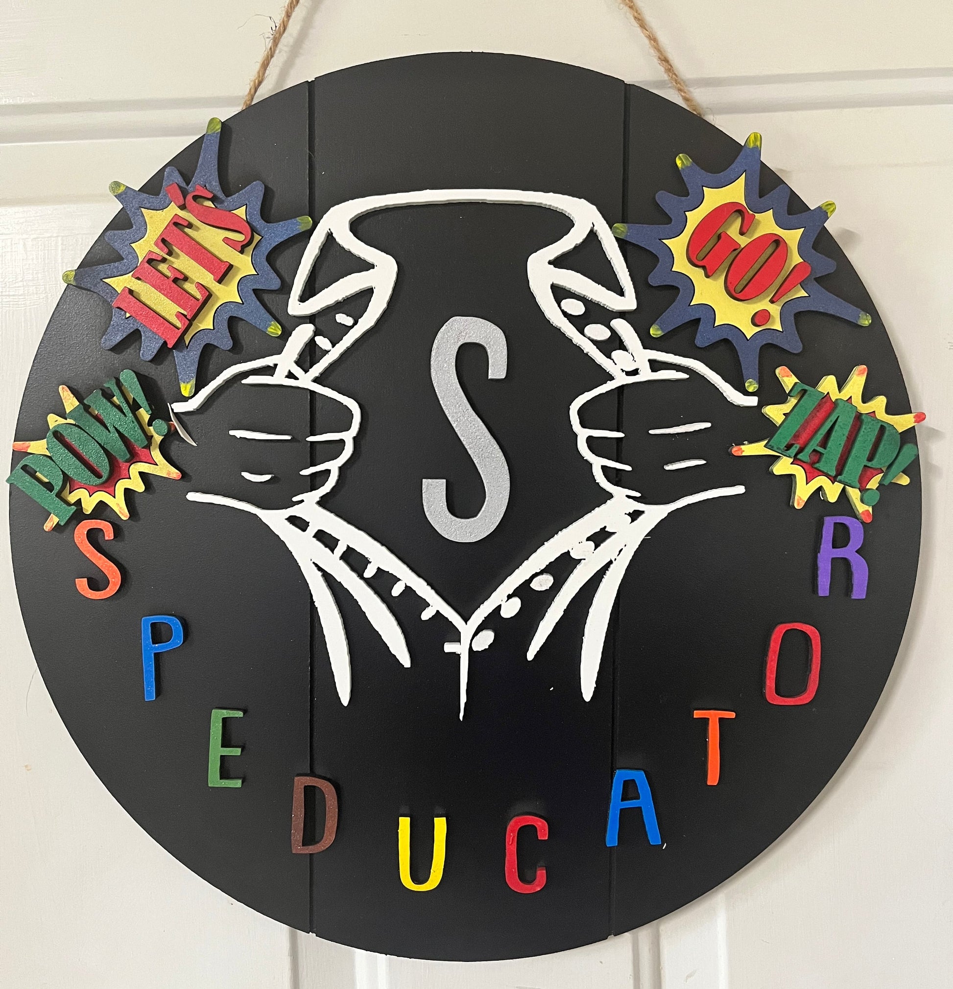 A circular black wall sign with a white line drawing of hands holding a banner, surrounded by colorful comic-style burst shapes and multicolored letters spelling a phrase, shown hanging against a white paneled wall.