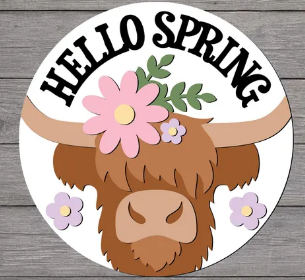 A circular spring-themed graphic featuring a brown highland cow illustration with a pink flower on its horn, surrounded by small purple flowers and green leaves. The words 'HELLO SPRING' are written in black arched lettering across the top, set against a gray wood-plank background.