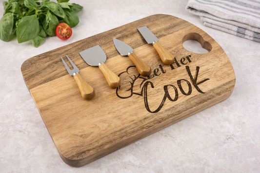 Wooden cutting board with cheese tools and 'Let Her Cook' engraving on a light background