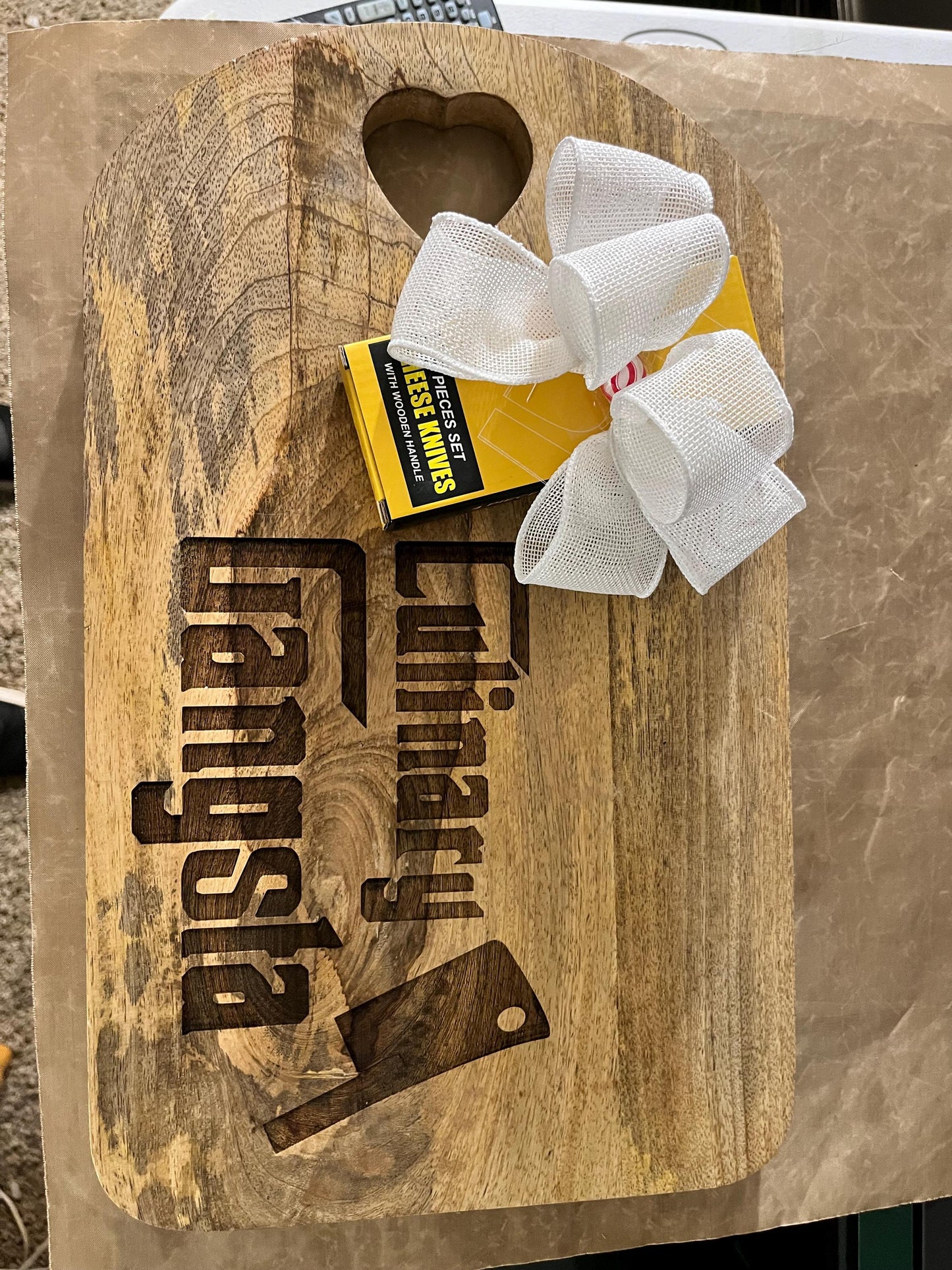 Wooden cutting board with 'Culinary Gangster' engraving, matches, and a bow on a wooden surface.