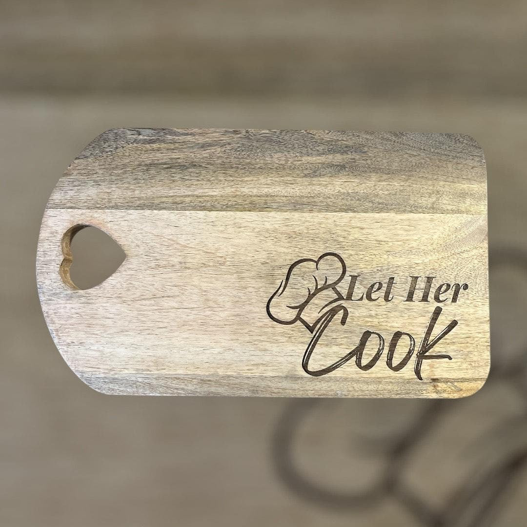 Wooden cutting board with 'Let Her Cook' engraving on a blurred background